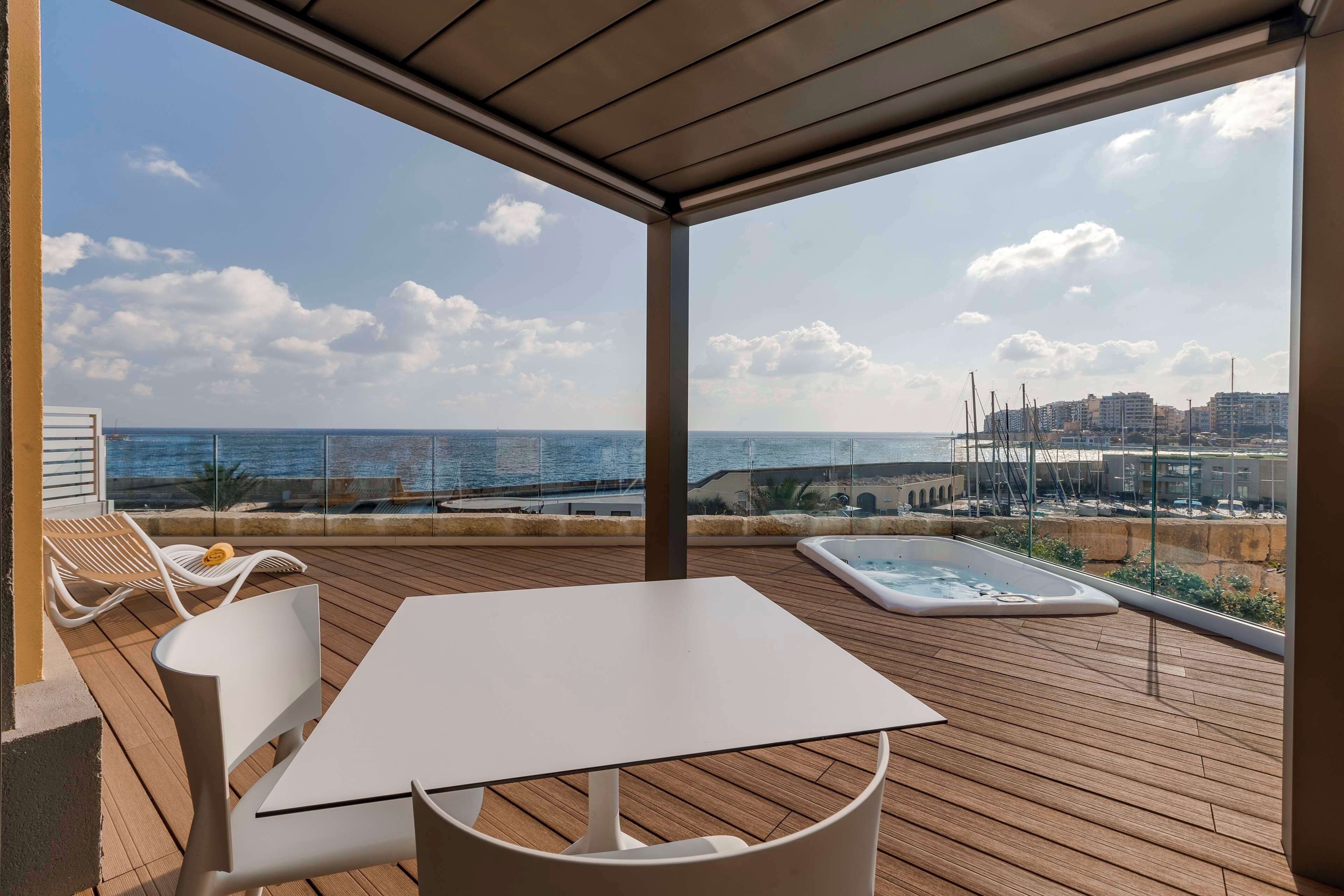 A wooden deck with a table and chairs, a hot tub, and a view of the sea and buildings.