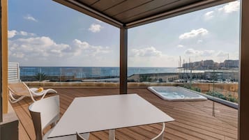 A wooden deck with a table and chairs, a hot tub, and a view of the sea and buildings.