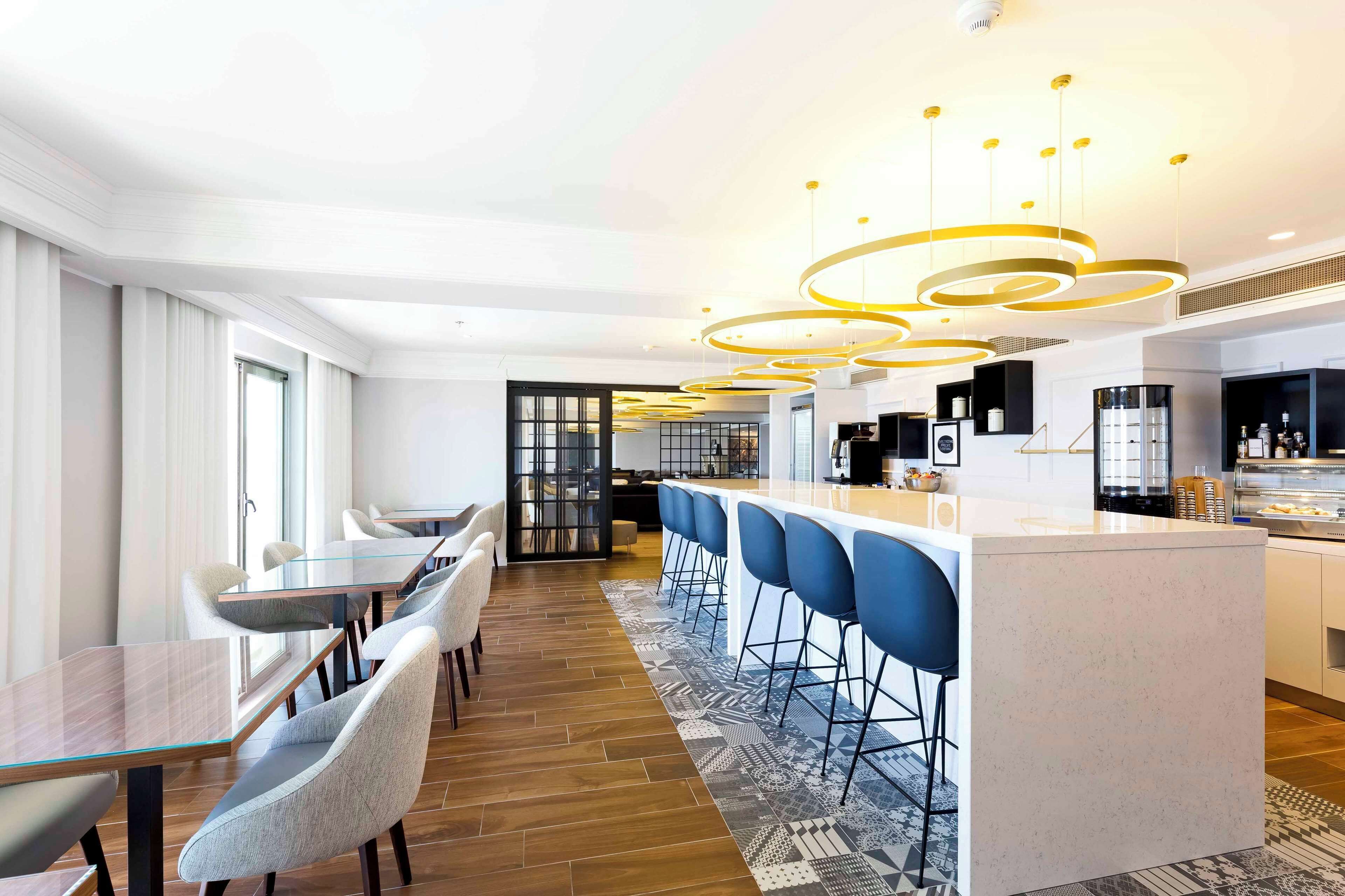 A modern hotel lobby with a long reception desk, bar area with blue stools, and a patterned rug.
