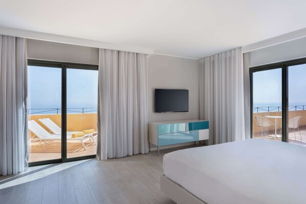 A hotel room with a large bed, a TV mounted on the wall, a sliding glass door leading to a balcony with a pool and lounge chairs, and a modern bathroom with a glass shower enclosure.