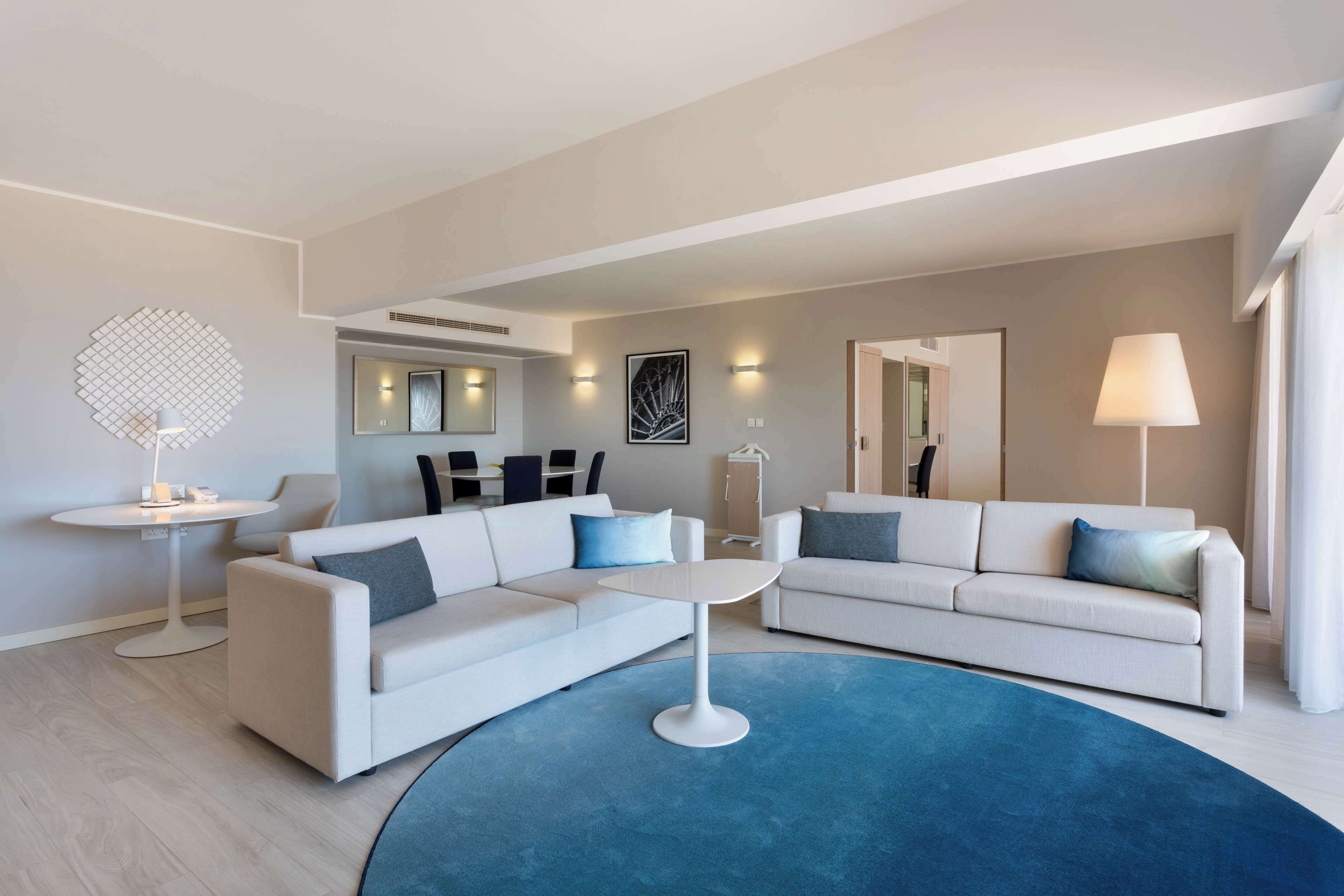 A modern living room with two white sofas, a round coffee table, and a blue rug. There are wall-mounted lights and a large mirror.