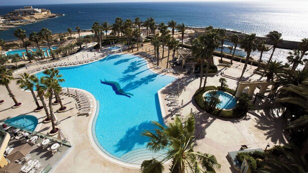 A resort with a large swimming pool, palm trees, and a view of the ocean. - Hilton Malta (St. Julian's)