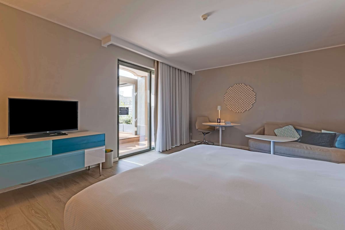 Une chambre d’hôtel moderne avec un grand lit, un téléviseur à écran plat posé sur une commode bleue et blanche, une petite table avec une lampe et un canapé avec des coussins.