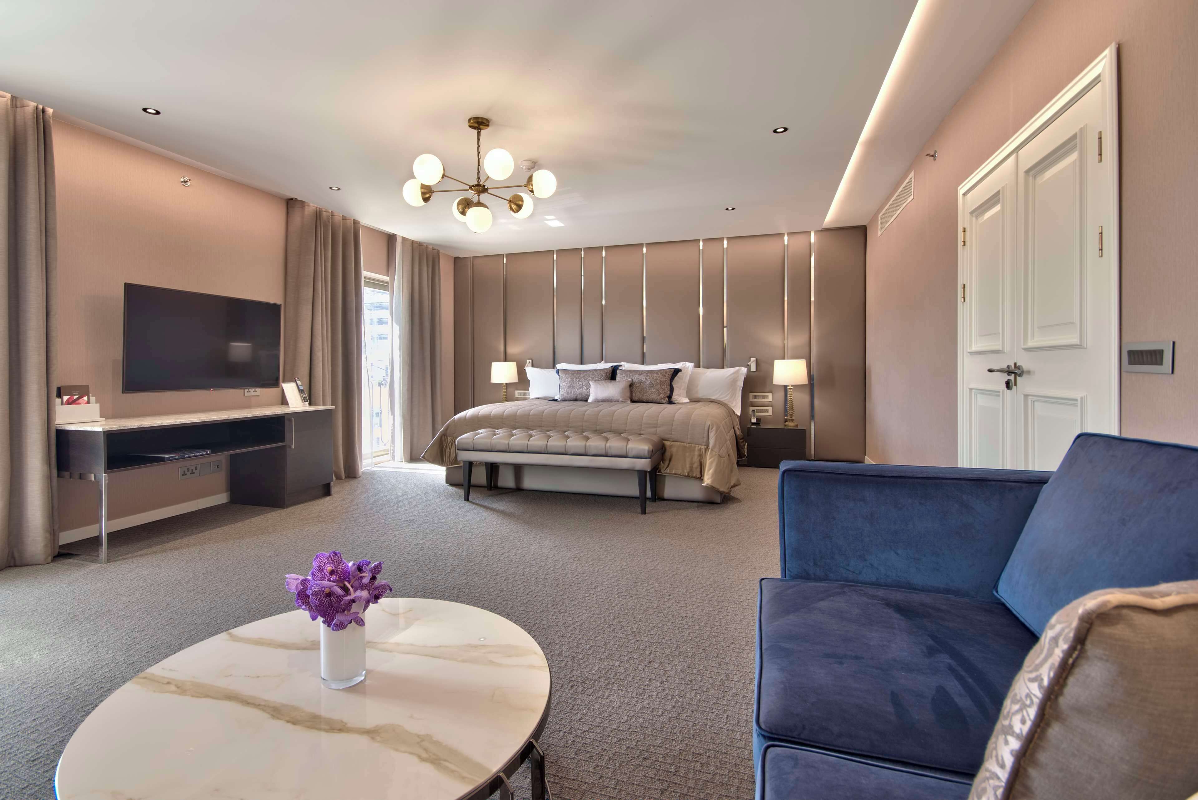 A modern hotel room with a large bed, a flat-screen TV, a sofa, and a marble coffee table with a vase of flowers.
