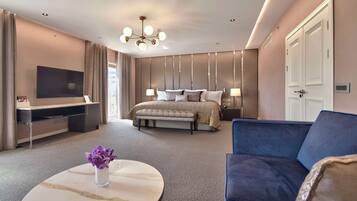 A modern hotel room with a large bed, a flat-screen TV, a sofa, and a marble coffee table with a vase of flowers.
