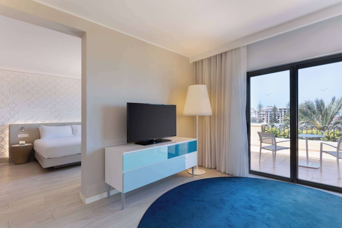Une chambre d’hôtel moderne avec un lit, un téléviseur posé sur une console blanche, une grande fenêtre donnant sur des palmiers et des immeubles, et une moquette bleue.