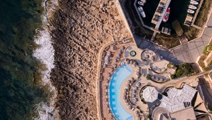 An aerial view of a coastal resort with a large swimming pool, beach area, and several boat docks. - Hilton Malta (St. Julian's)