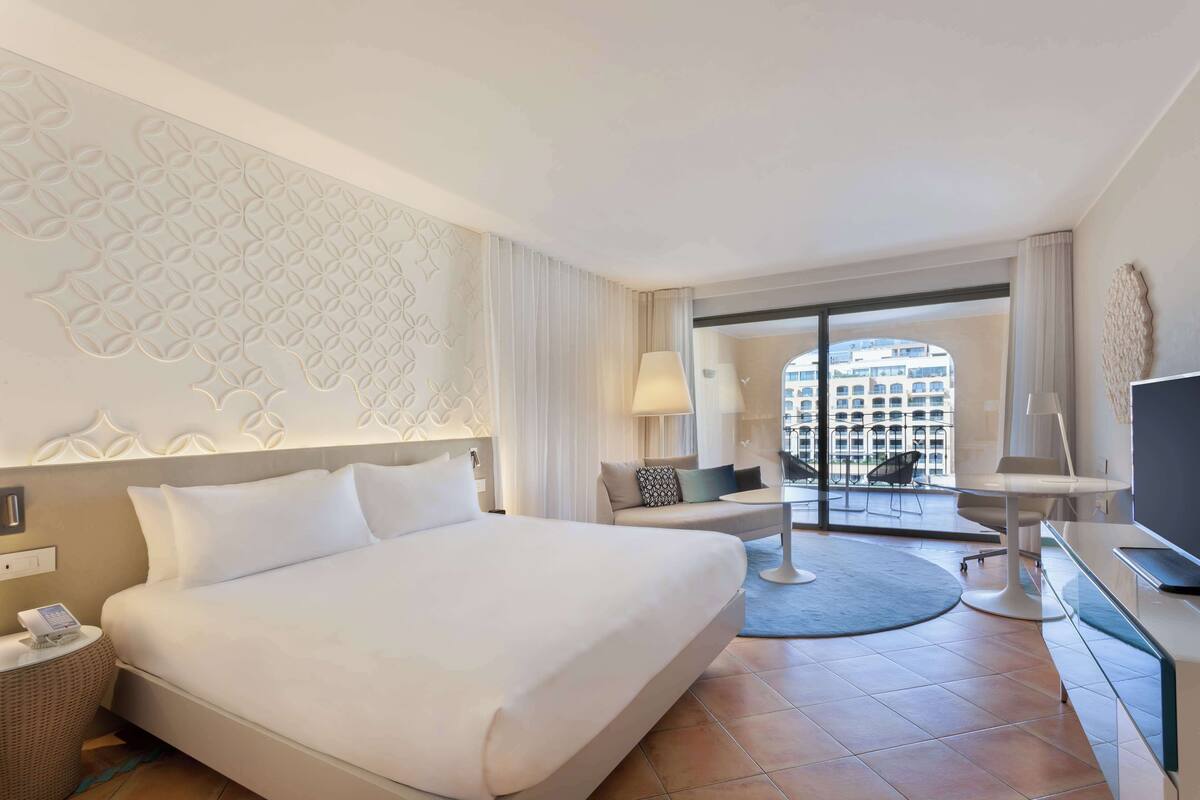Une chambre d’hôtel moderne dotée d’un grand lit, d’un bureau avec un ordinateur, d’une chaise et offrant une vue sur la ville grâce à une grande fenêtre.