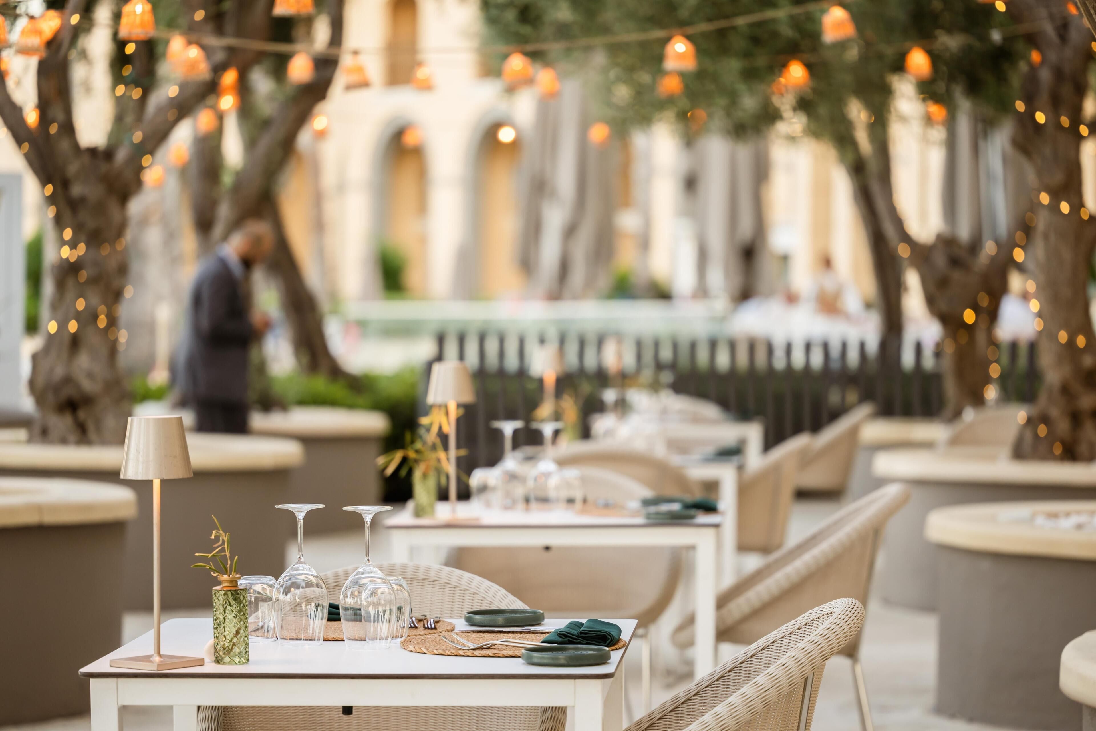 Un área de comedor al aire libre con mobiliario de mimbre, mesas dispuestas con platos, vasos y un jarrón con flores.