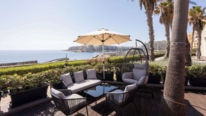 An outdoor seating area with a sofa, chairs, and a table, overlooking a coastal view with palm trees and buildings in the background. - Hilton Malta (St. Julian's)