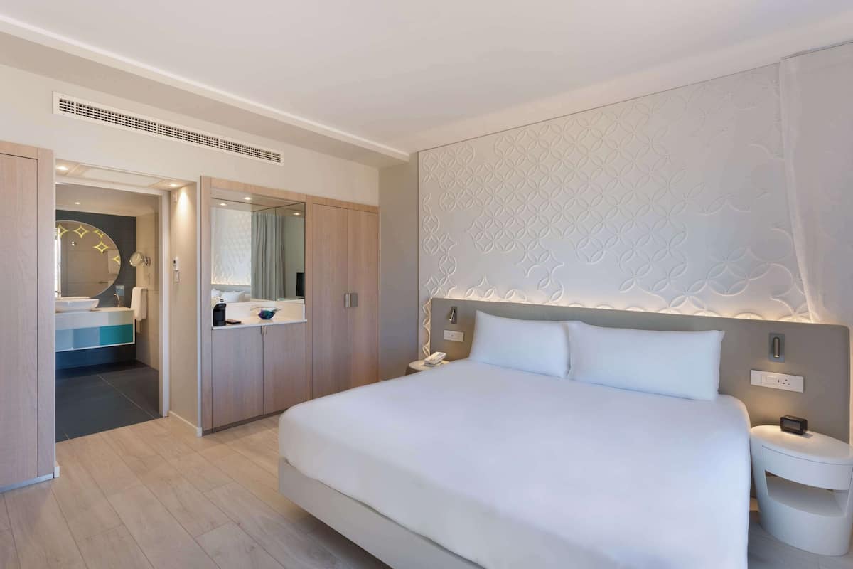 Une chambre d’hôtel moderne avec un grand lit, une table de chevet avec un téléphone et une radio, et une salle de bain équipée d’un miroir, d’un lavabo et d’un porte-serviettes.