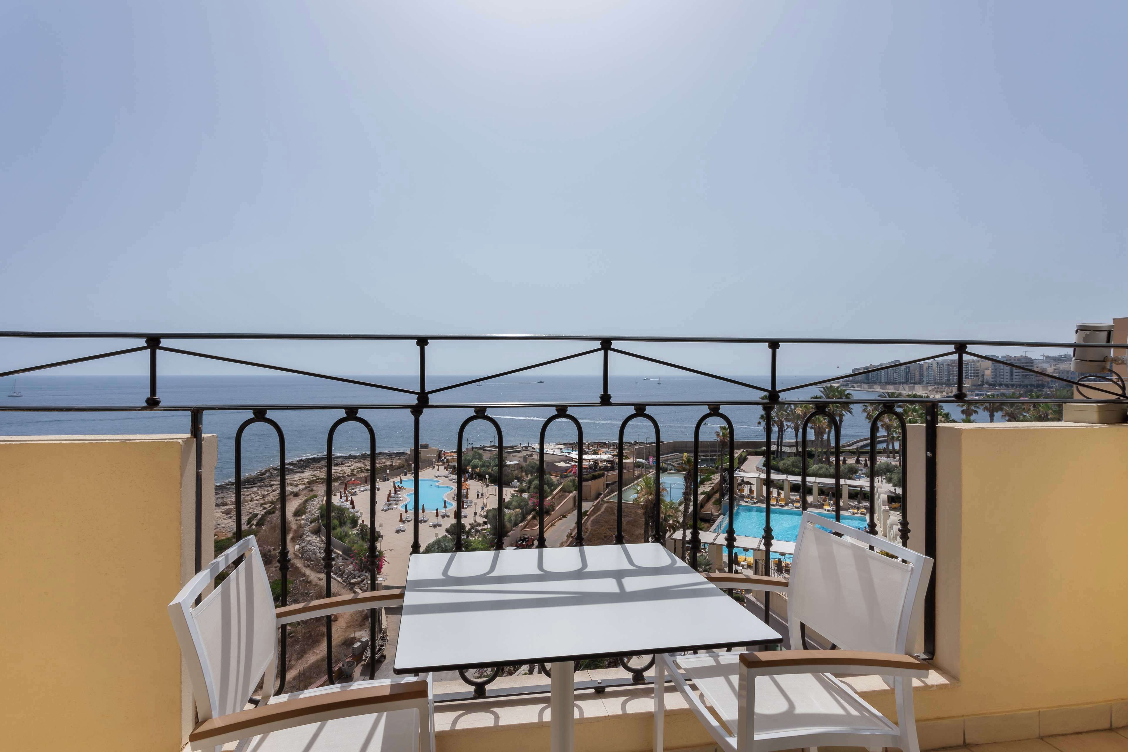 A balcony with a table and chairs, overlooking a coastal area with a swimming pool and buildings in the background.