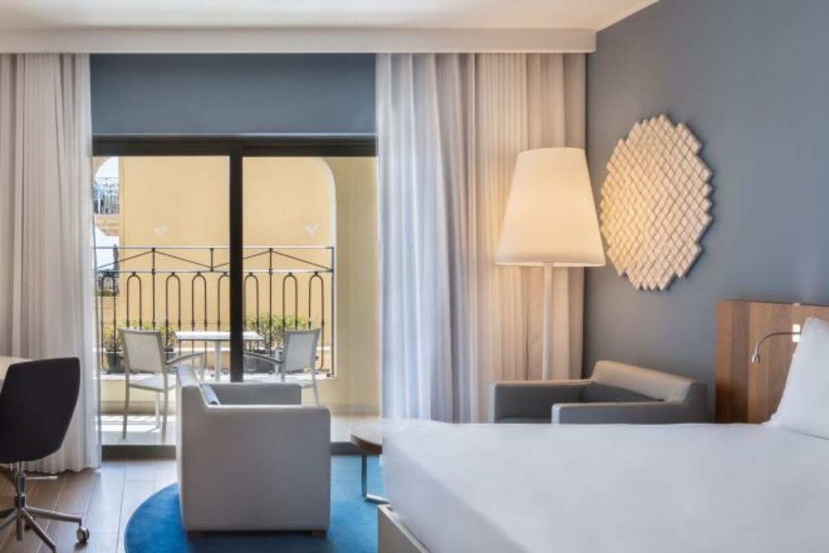 Habitación de hotel con cama, dos sillones, un escritorio y un balcón con mobiliario exterior.
