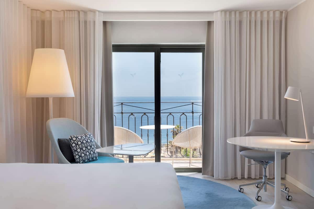 Une chambre d’hôtel avec un lit, deux chaises, une petite table, une lampe et une vue sur la mer depuis un balcon doté de rideaux.