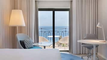Habitación de hotel con cama, dos sillas, una mesita, una lámpara y vista al mar desde un balcón con cortinas.