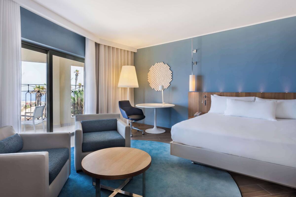 Une chambre d’hôtel comprenant un lit, deux fauteuils, une petite table, une lampe et une vue sur l’extérieur.