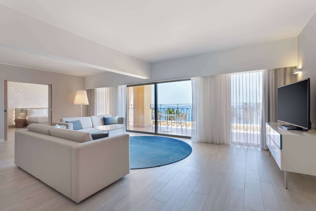 Une chambre d’hôtel moderne dotée d’un grand canapé, d’un téléviseur à écran plat, d’un balcon avec vue sur la mer et d’une grande fenêtre.