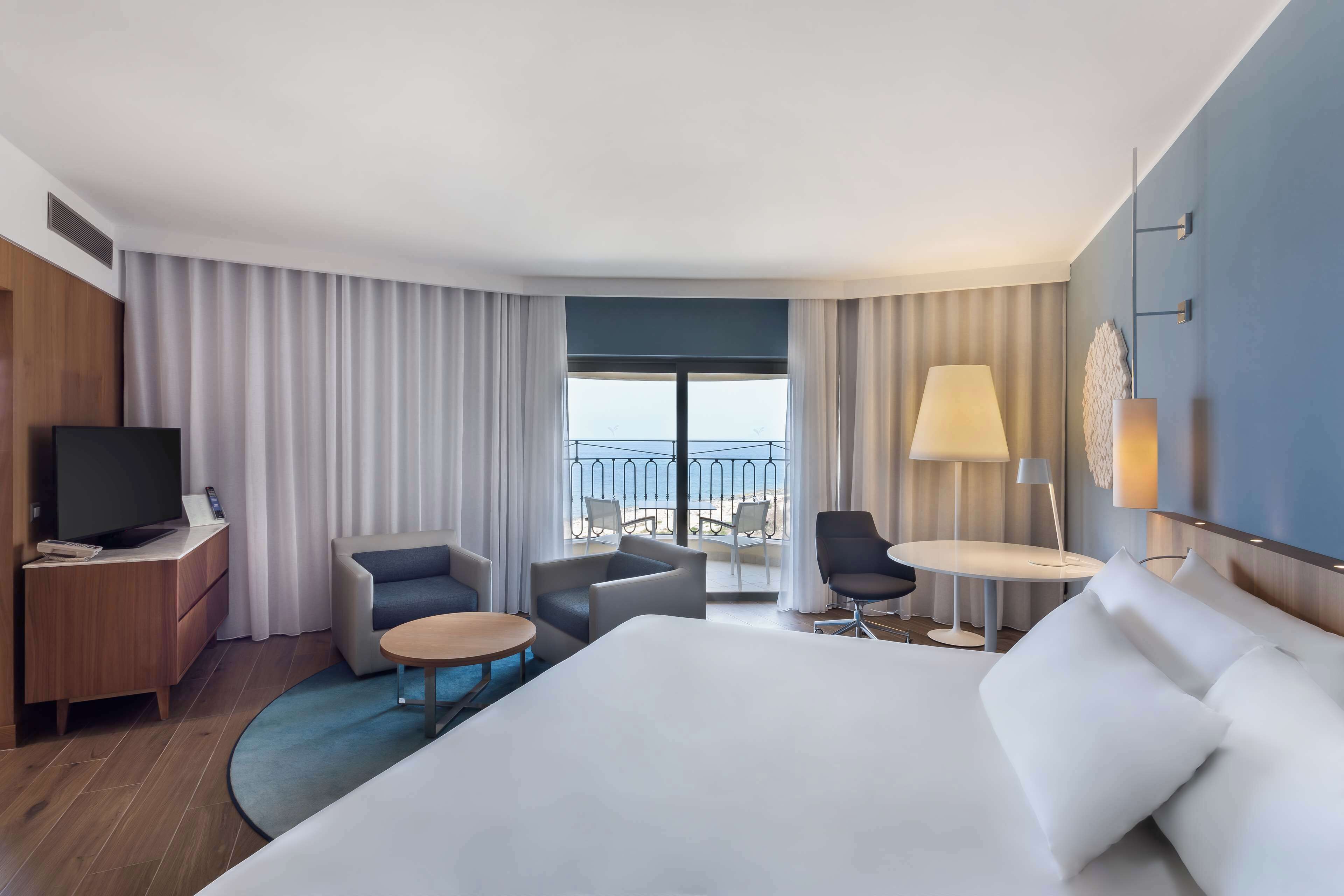 A hotel room with a large bed, a TV, a desk, and a balcony with a view.