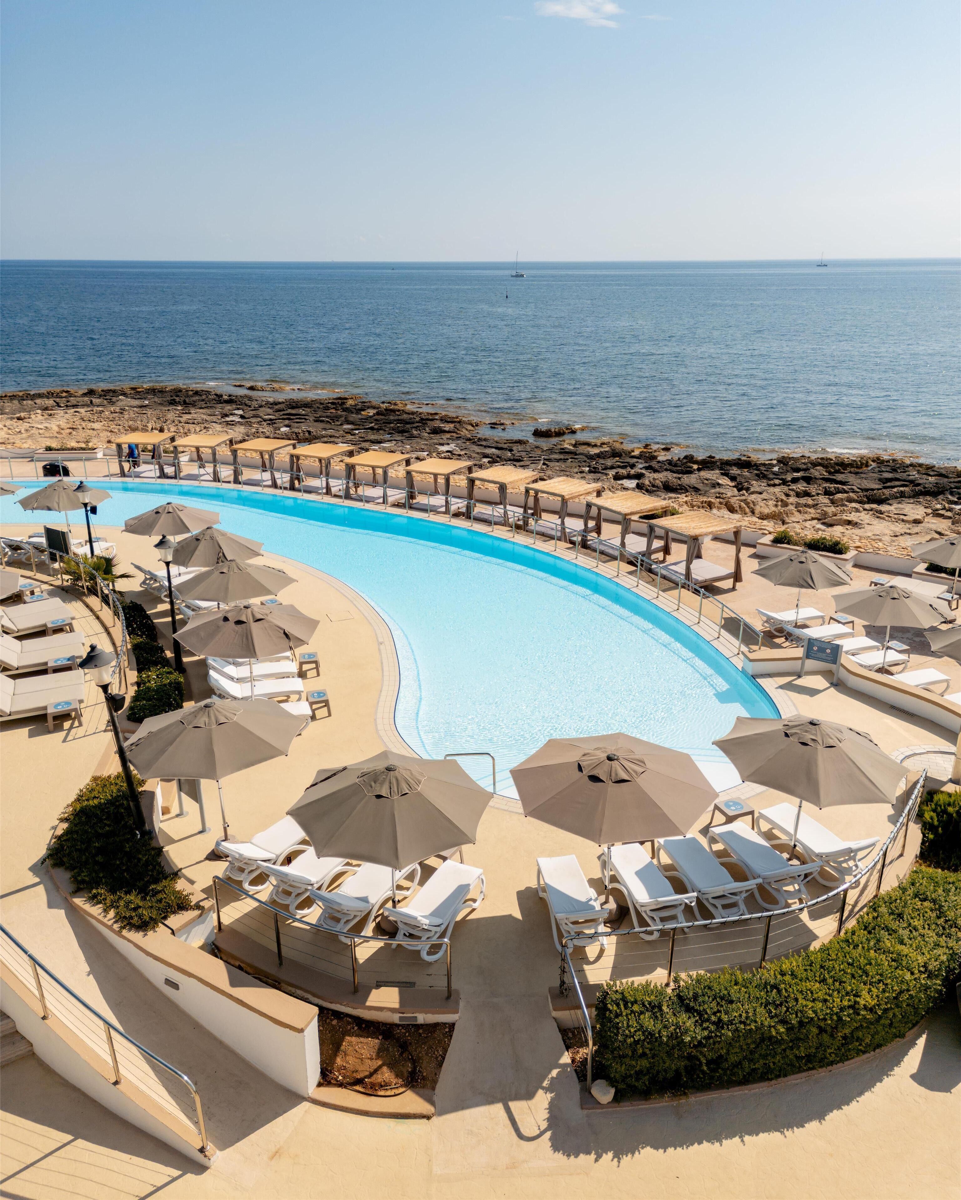 Una piscina con sillones de playa y sombrillas, una costa rocosa y el mar al fondo.