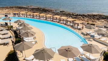 Una piscina con sillones de playa y sombrillas, una costa rocosa y el mar al fondo.