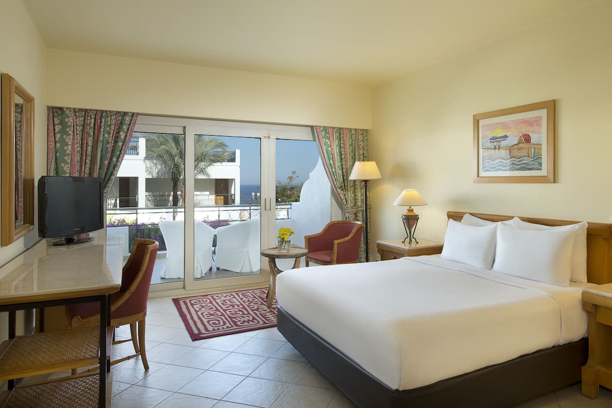 A hotel room with a bed, two chairs, a desk, a television, and a balcony with curtains.