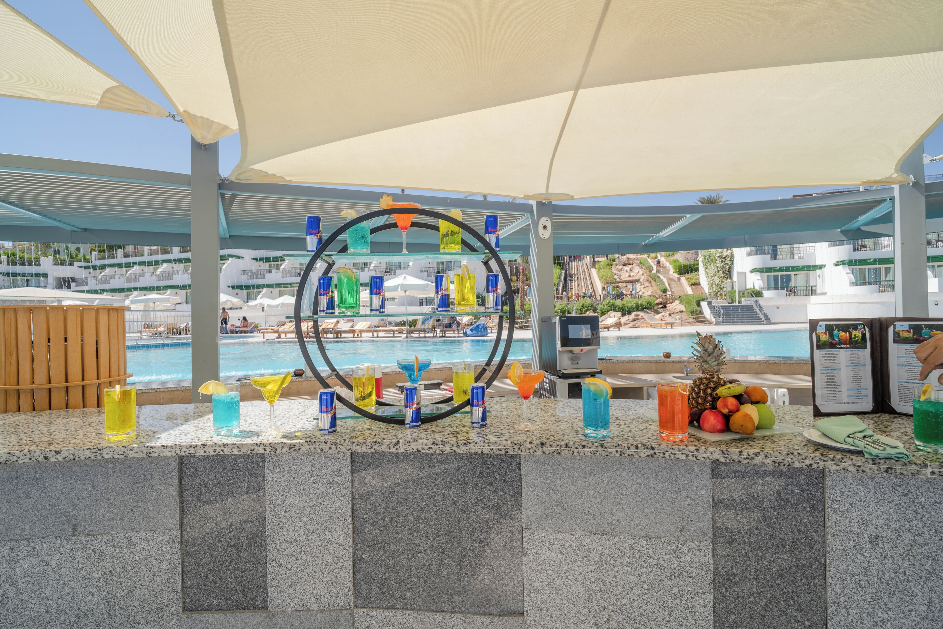 Zona de bar junto a la piscina, con un expositor de carta de bebidas, una variedad de refrescos y vistas a la piscina y los edificios del resort.