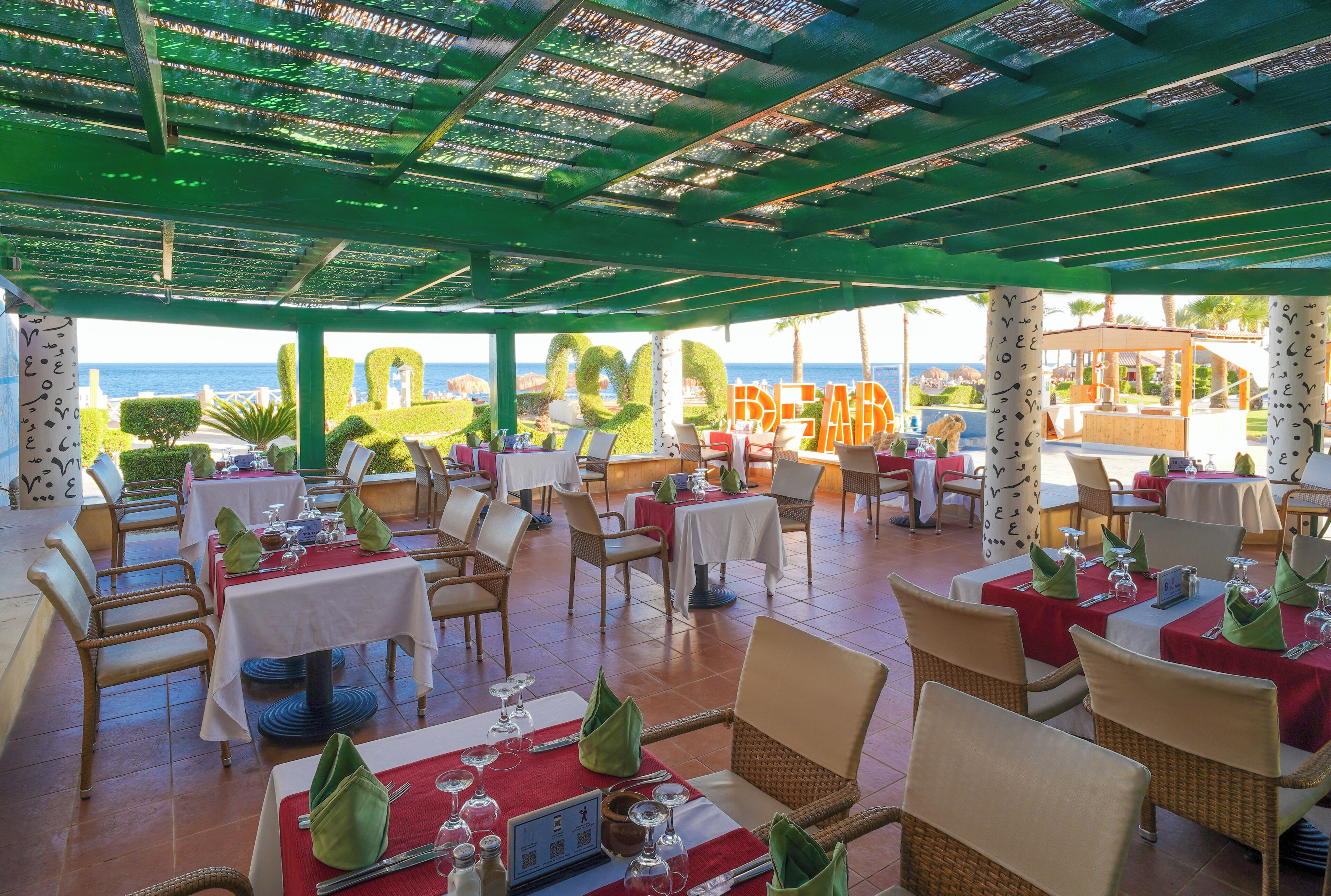 Un restaurante al aire libre con mesas y sillas, vista al mar y una zona cubierta con techo verde.