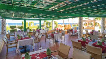 Un restaurante al aire libre con mesas y sillas, vista al mar y una zona cubierta con techo verde.
