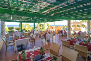 An outdoor restaurant with tables and chairs, a view of the sea, and a covered area with a green roof.