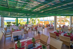 An outdoor restaurant with tables and chairs, a view of the sea, and a covered area with a green roof.