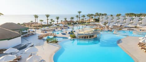 A resort pool area with multiple swimming pools, sun loungers, umbrellas, and a view of the ocean.