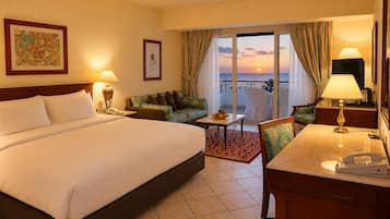 Habitación de hotel con una cama grande, un escritorio con teléfono, una silla, una mesita con fruta y vista al atardecer a través de las puertas del balcón.
