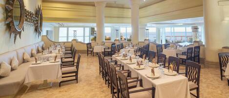 A spacious dining area with multiple tables and chairs, large windows offering a view, and a decorative mirror on the wall.