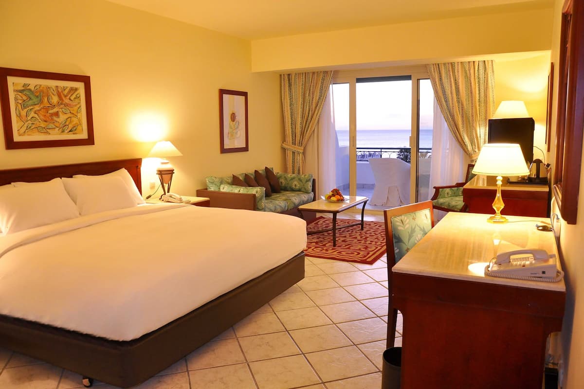 A hotel room with a large bed, a desk with a telephone, a chair, a small table with fruits, and a view of the ocean through the balcony doors.