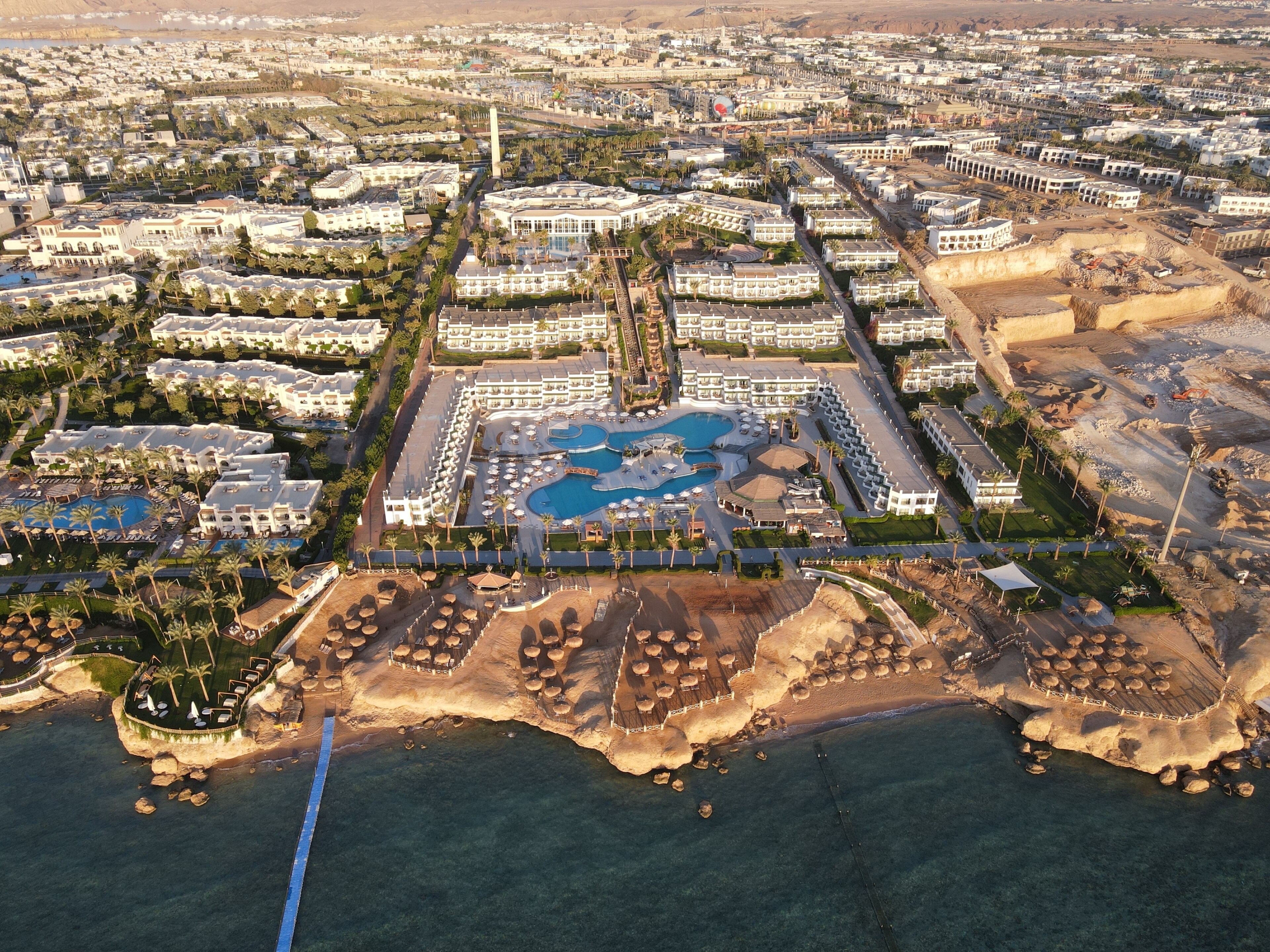 Vue aérienne d’un complexe hôtelier comprenant plusieurs bâtiments, un espace piscine central et une plage avec des transats.