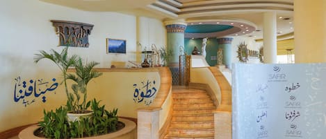 A grand lobby with a marble staircase, decorative columns, and a reception desk with multilingual signage.