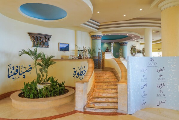 A grand lobby with a marble staircase, decorative columns, and a reception desk with multilingual signage.