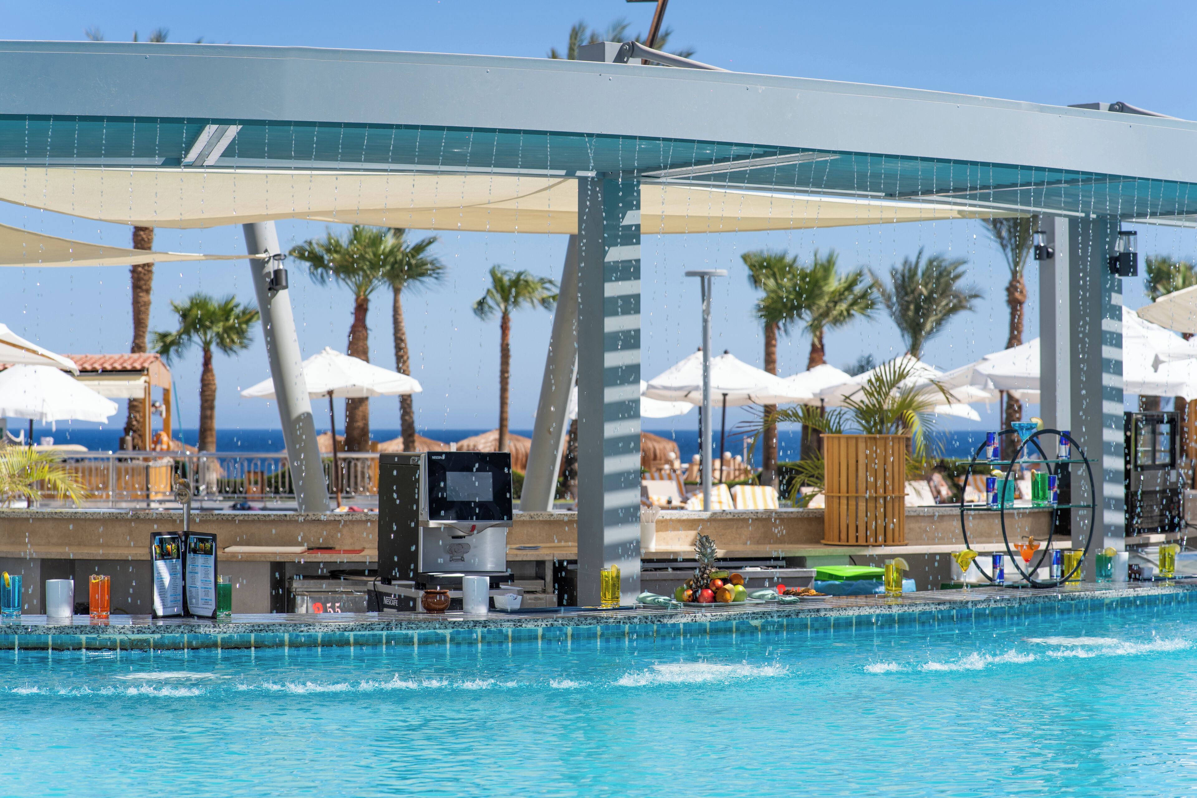 Zona de piscina con bar, sombrillas y palmeras.
