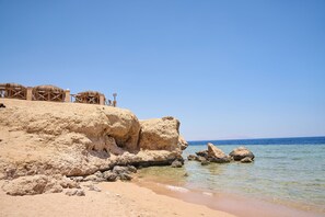 A beach with a rocky cliff, clear blue sky, and calm sea. - Safir Sharm Waterfalls Resort (Sharm El Sheikh)