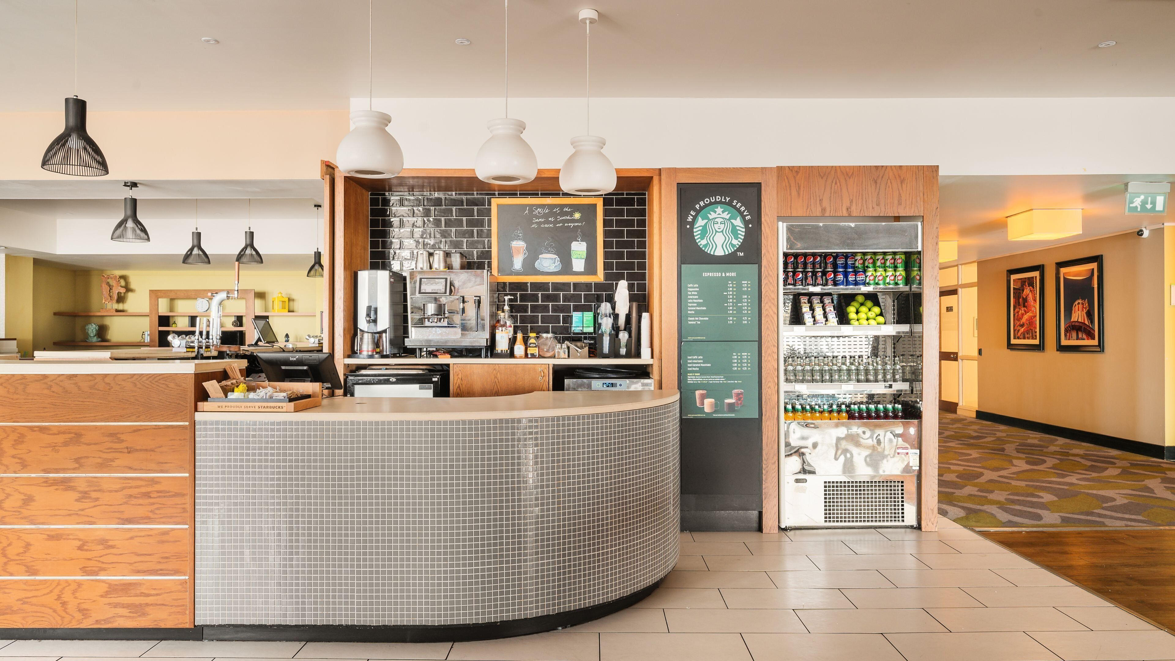 Interior moderno de una cafetería con un mostrador curvo, máquinas de espresso, una pizarra de menú y un dispensador de bebidas.