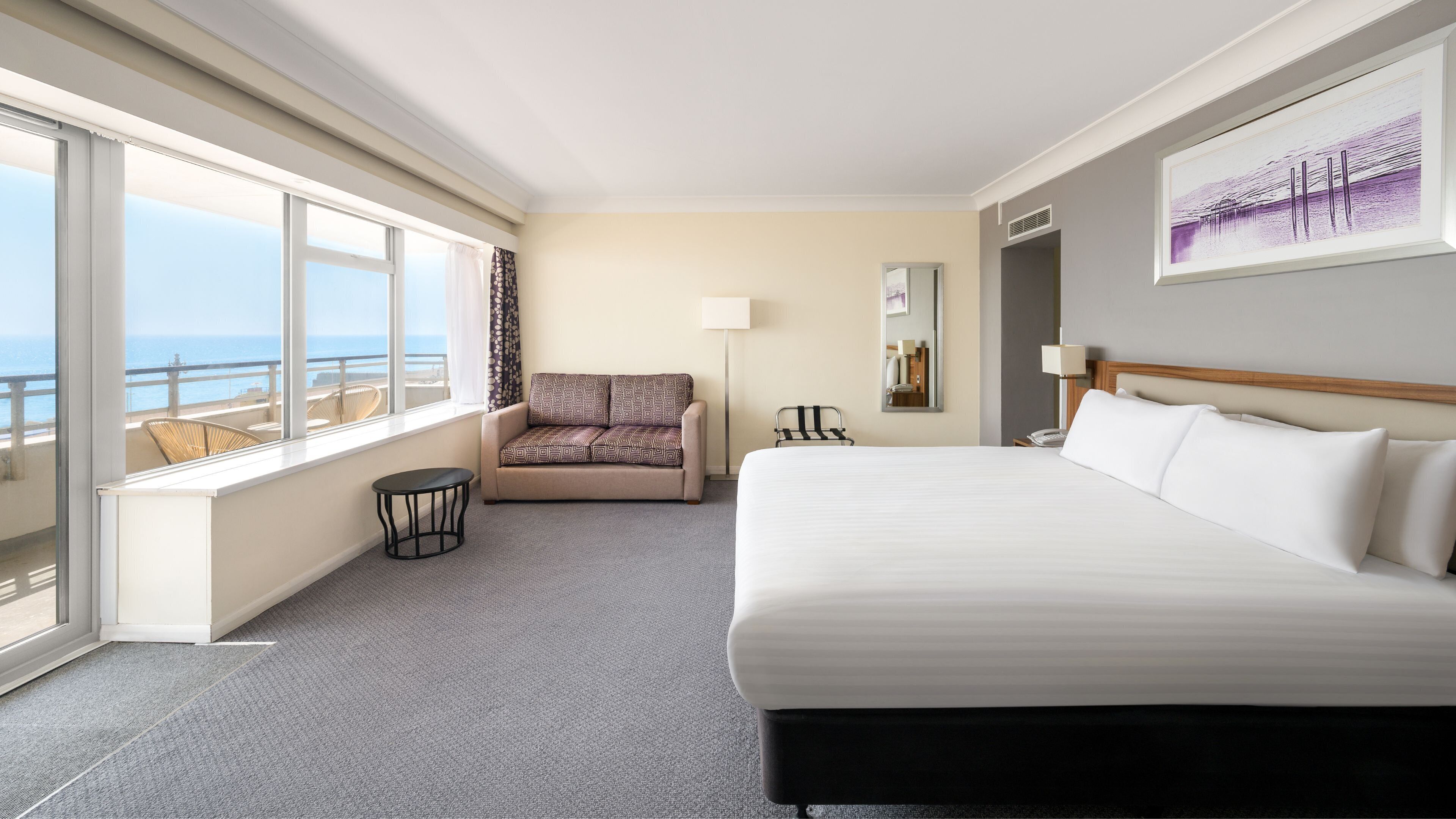 A hotel room with a large bed, a sofa, a chair, a small table, a lamp, and a view of the ocean.
