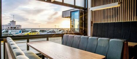 A modern interior with a wooden table and cushioned seating, large windows offering a view of the sea and parked vehicles.
