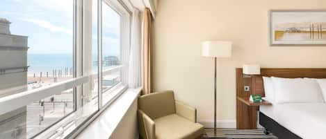 A hotel room with a large window offering a view of the beach and pier, a bed with white linens, a chair, a bedside table with a lamp, and a framed picture on the wall.
