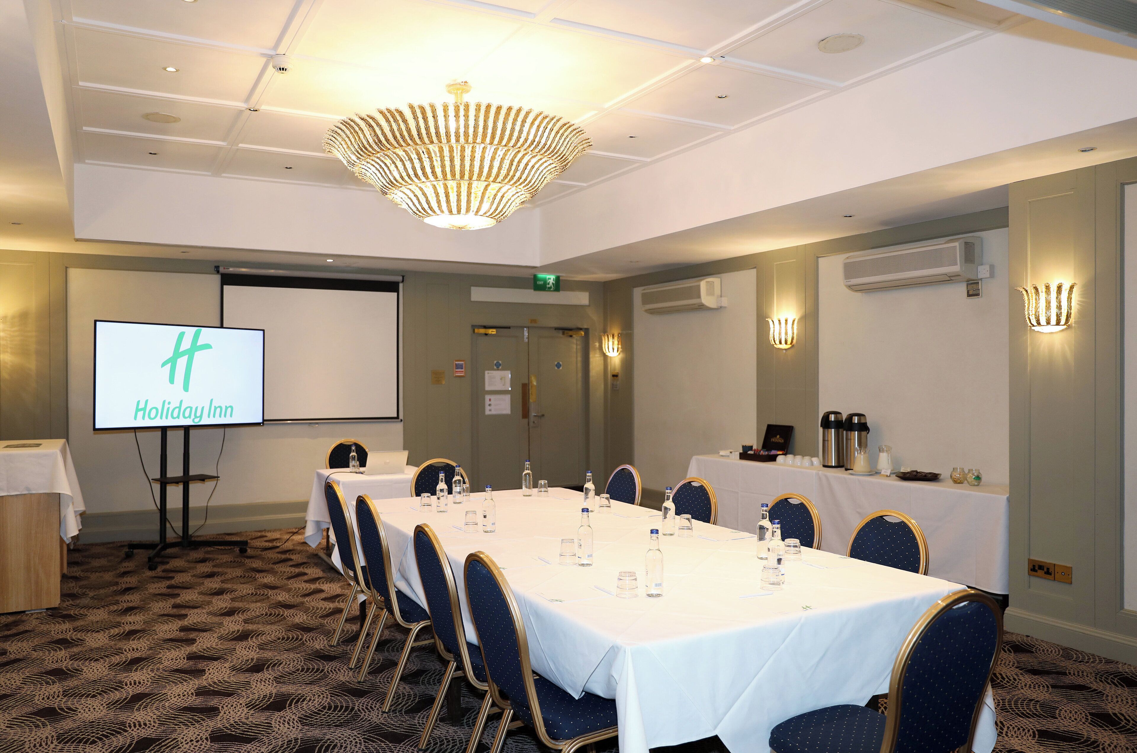 A conference room with a long table, chairs, a projector screen displaying "Holiday Inn", and a chandelier.