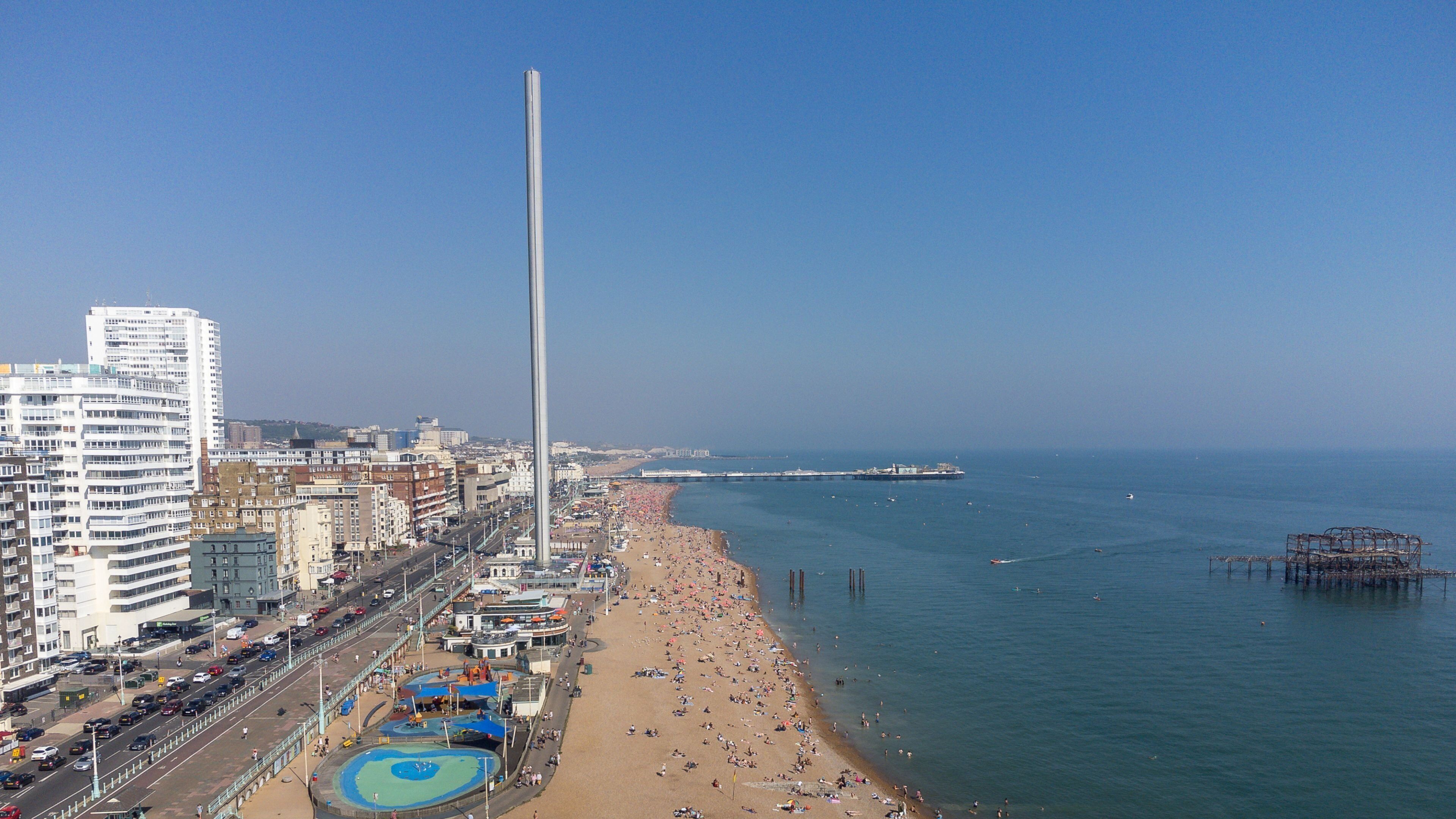 Paesaggio costiero urbano con un imponente monumento, una spiaggia affollata e un molo che si estende verso il mare.