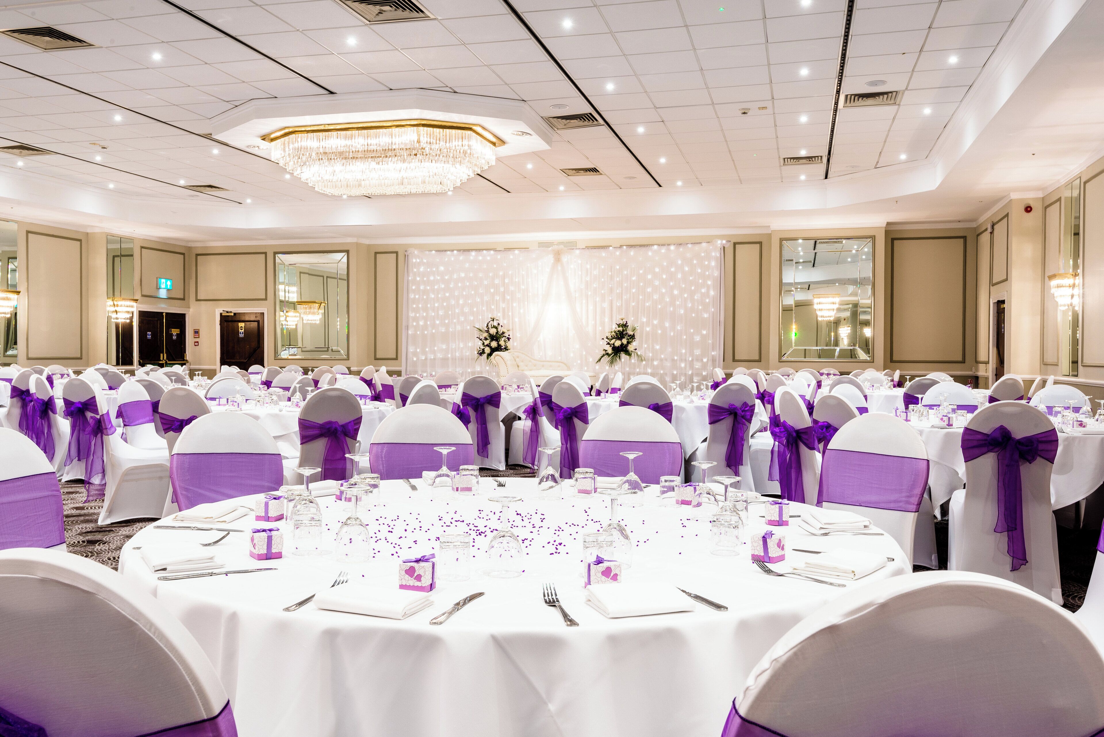 Un salón de banquetes con mesas redondas, manteles blancos y cintas moradas en las sillas.