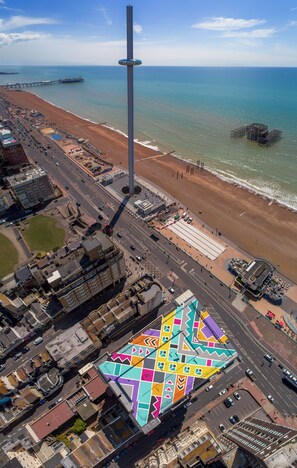 An aerial view of a coastal city with a prominent tower, a colorful building with geometric patterns, and a beach with a pier.