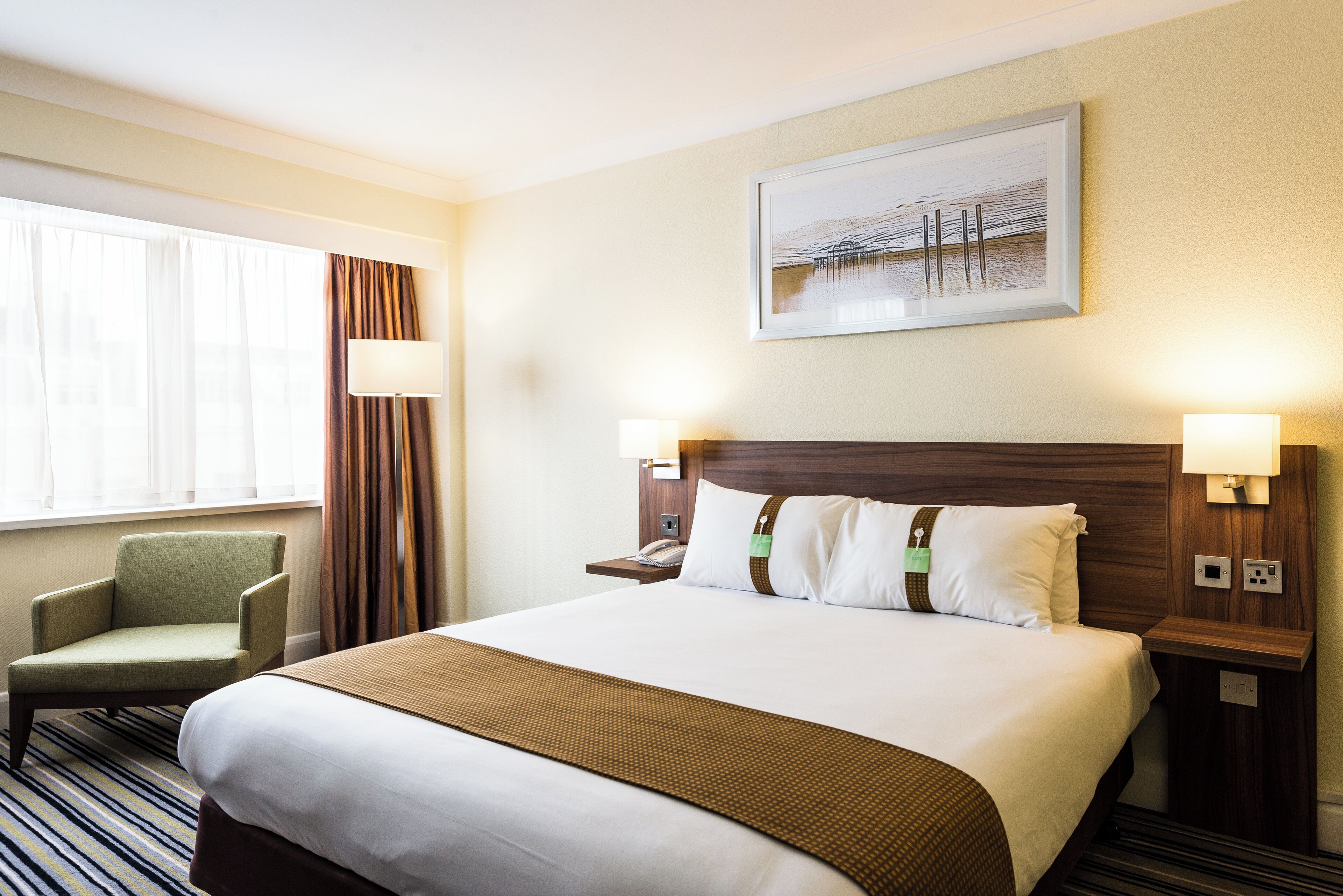 Une chambre d’hôtel avec un grand lit, une chaise, une table de chevet, une lampe et un tableau au mur.