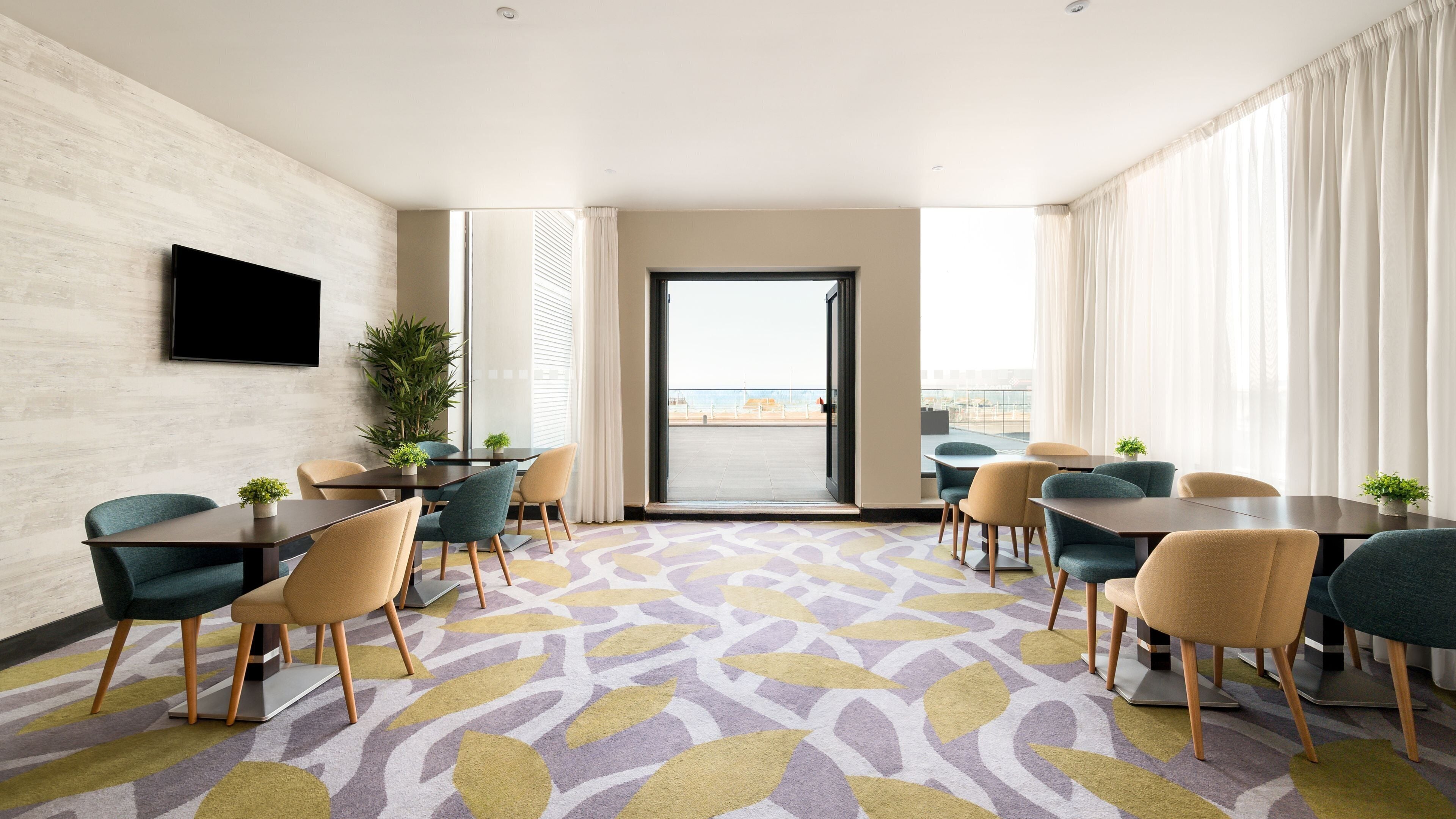 A hotel room with a large window, a flat-screen TV mounted on the wall, and a seating area with tables and chairs.