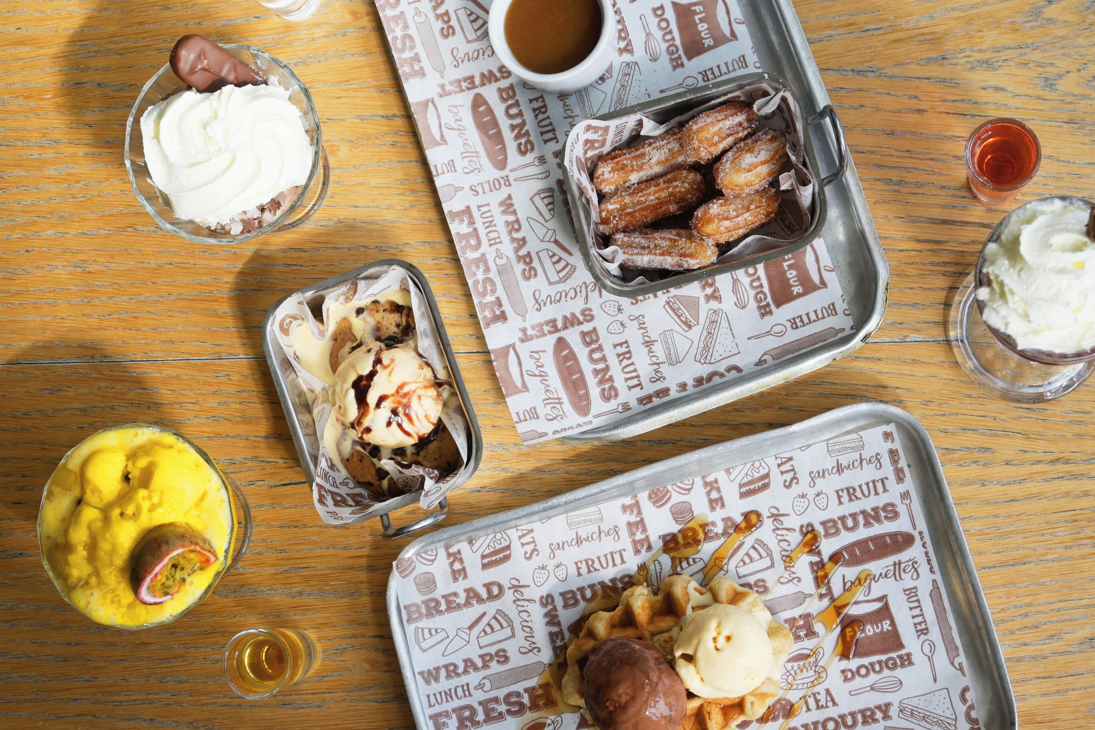 Una mesa de madera con diversos alimentos, incluyendo churros, helado y una bebida.
